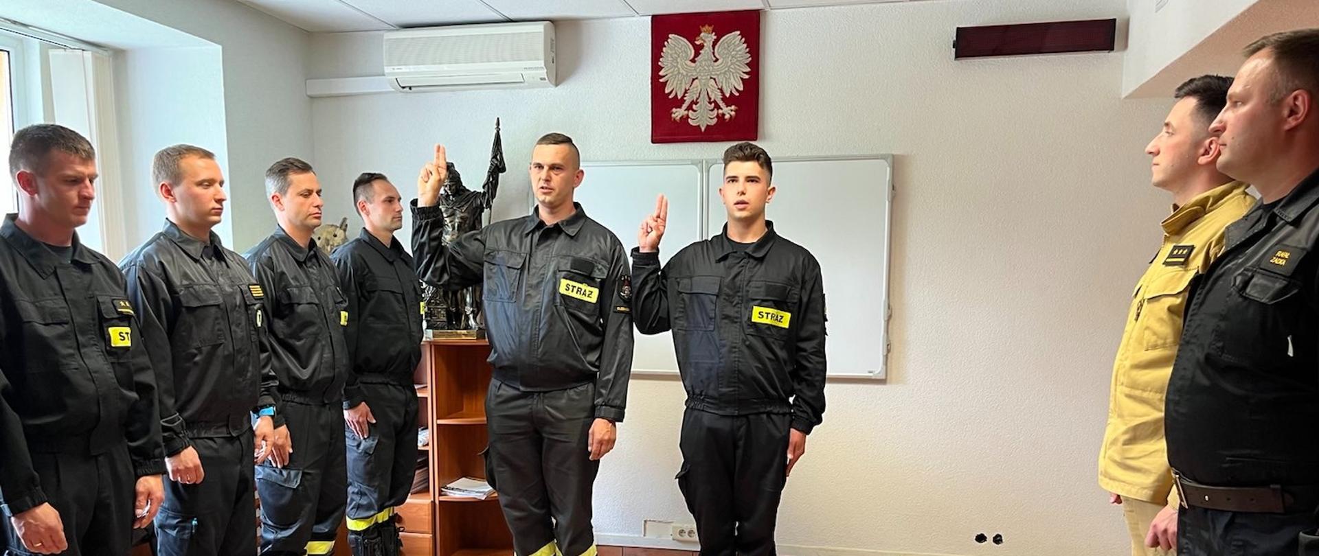 Na zdjęciu widać strażaków stojących w szyku. Na środku dwóch strażaków stoi z uniesioną prawą ręką zgiętą w łokciu - składają ślubowanie Komendantowi Powiatowemu PSP w Oleśnicy. 
