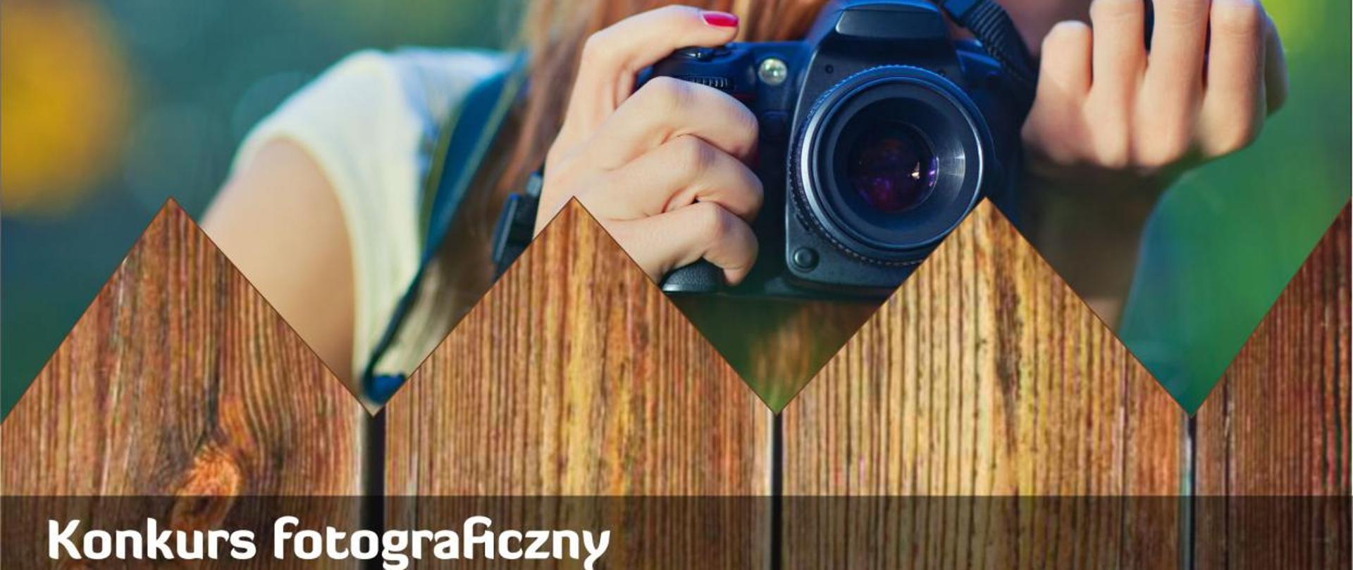 Na zdjęciu widać drewniany płot, za płotem stoi kobieta na tle roślinności robiąca zdjęcie czarnym aparatem. Na dole zdjęcia znajduje się biały napis "Konkurs fotograficzny".