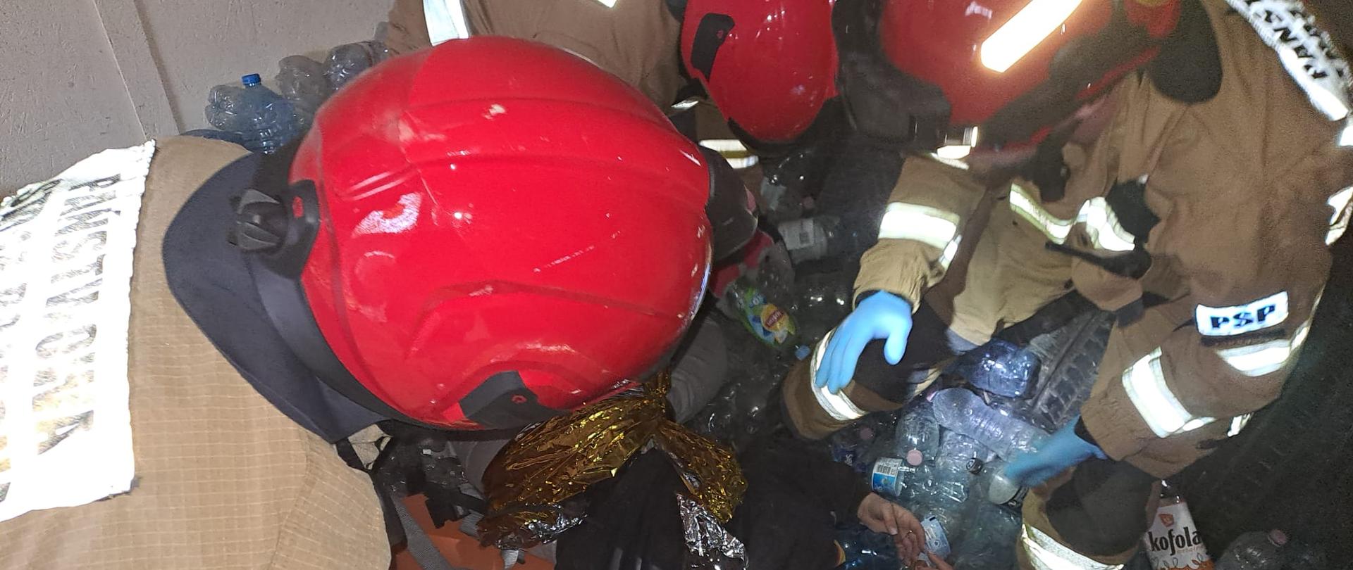 Strażacy PSP w ubraniach specjalnych i czerwonych hełmach prowadzą działania poszukiwawczo‑ratownicze w ciasnej przestrzeni wypełnionej plastikowymi butelkami. Pochylają się nad poszkodowanym, udzielając mu pomocy. Ujęcie z ćwiczeń realizowanych podczas szkolenia podstawowego KSRG.
