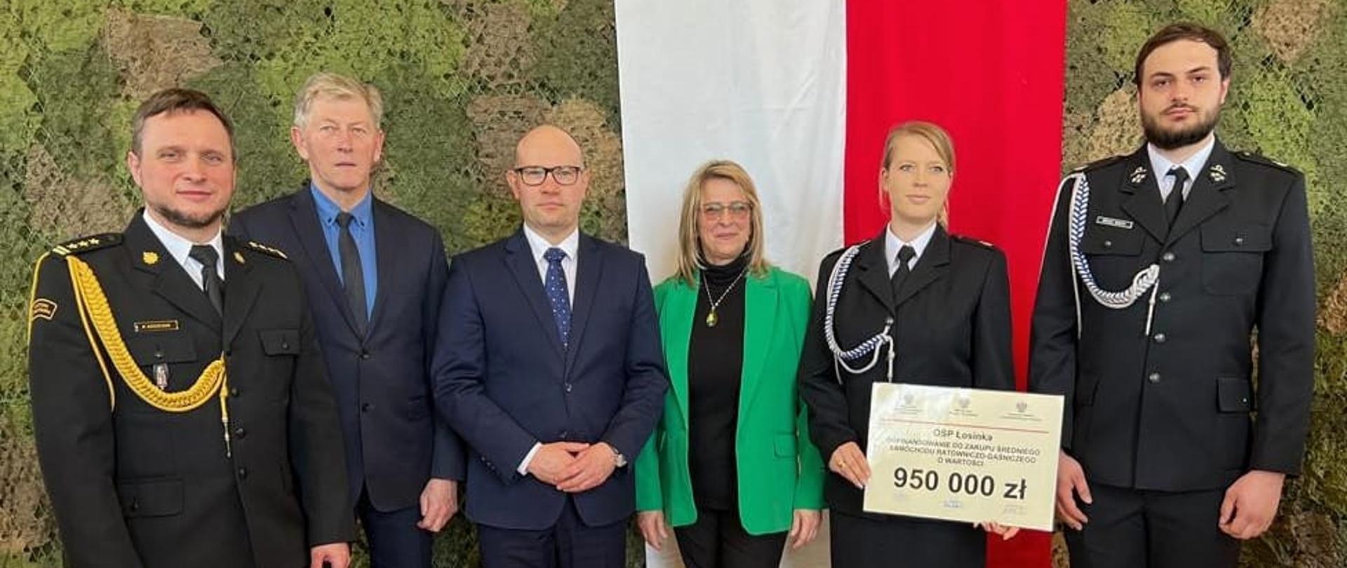 W pomieszczeniu w rzędzie na tle flagi stoi kilka druhen i druhów w mundurach oraz osoby w cywilnym ubraniu.