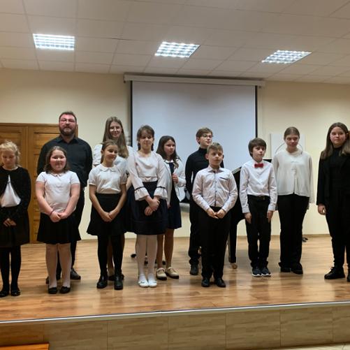 Na scenie wykonawcy koncertu z klasy fortepianu p. Piotra Wilka wraz z nauczycielem. Wszyscy stoją w dwóch rzędach ubrani na galowo. W tle fragmenty czarnego fortepianu. 