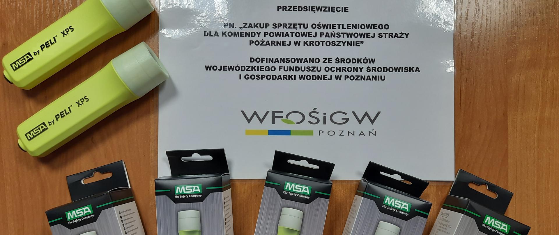 Zdjęcie przedstawia latarki strażacki koloru zielonego. Dwie latarki leżą bez opakowania gotowe do użycia a pięć latarek zapakowane w oryginalne opakowania. Na latarkach widoczny jest napis MSA by PELI XPS. Na zdjęciu widoczne jest kartka z napisem "Przedsięwzięcie pn. Zakup sprzętu oświetleniowego dla Komendy Powiatowej Państwowej Straży Pożarnej w Krotoszynie dofinansowano ze środków Wojewódzkiego Funduszu Ochrony Środowiska i Gospodarki Wodnej w Poznaniu.