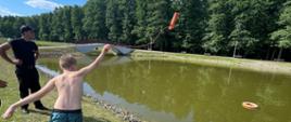 Na zdjęciu widać chłopca który z brzegu zbiornika wodnego przy asyście strażaków rzuca do wody rzutkę ratowniczą. Próbuje trafić do celu jakim jest koło ratownicze pływające po wodzie