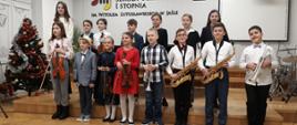 Na zdjęciu widnieje grupa uczniów, ustawionych w dwóch rzędach na scenie auli PSM I stopnia w Jaśle. Niektórzy z uczniów trzymają w rękach skrzypce, saksofony i trąbki. Przeważająca kolorystyka zdjęcia: brązowo-czarno-biała.