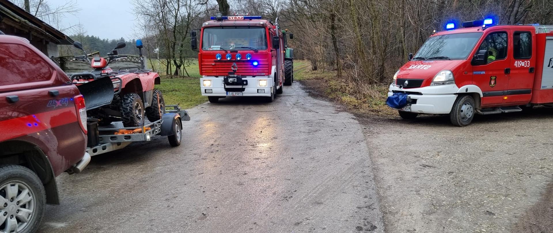 Zdjęcie przedstawia trzy samochody ratowniczo-gaśnicze, w tym jeden lekki samochód rozpoznawczo-ratowniczy z przyczepą, na której znajduje się czterokołowiec typu quad. Zdjęcie zrobione na skraju lasu.
