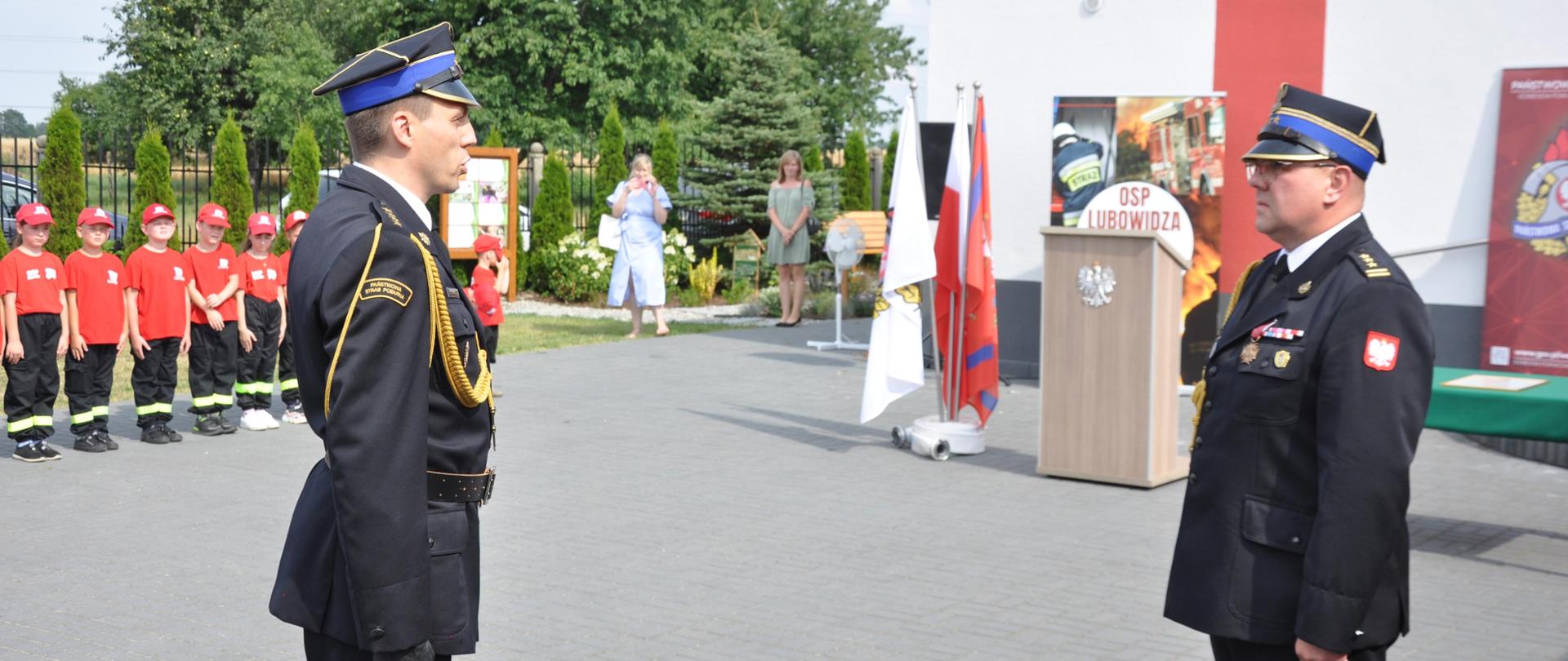 Dowódca uroczystości składa meldunek Zastępcy Łódzkiego Komendanta Wojewódzkiego Państwowej Straży Pożarnej st. bryg. Radosławowi Pełce z okazji włączenia jednostki Ochotniczej Straży Pożarnej w Lubowidzy do Krajowego Systemu Ratowniczo-Gaśniczego. Uroczystość ma miejsce na placu przed jednostką. Na drugim planie stoi mównica i stojak flagowy. W tle widać część budynku strażnicy. Po lewej stronie w pododdziale stoi młodzieżowa drużyna pożarnicza ubrana w czerwone koszulki i czapki. Rzecz dzieje się na placu jednostki osp lubowidza