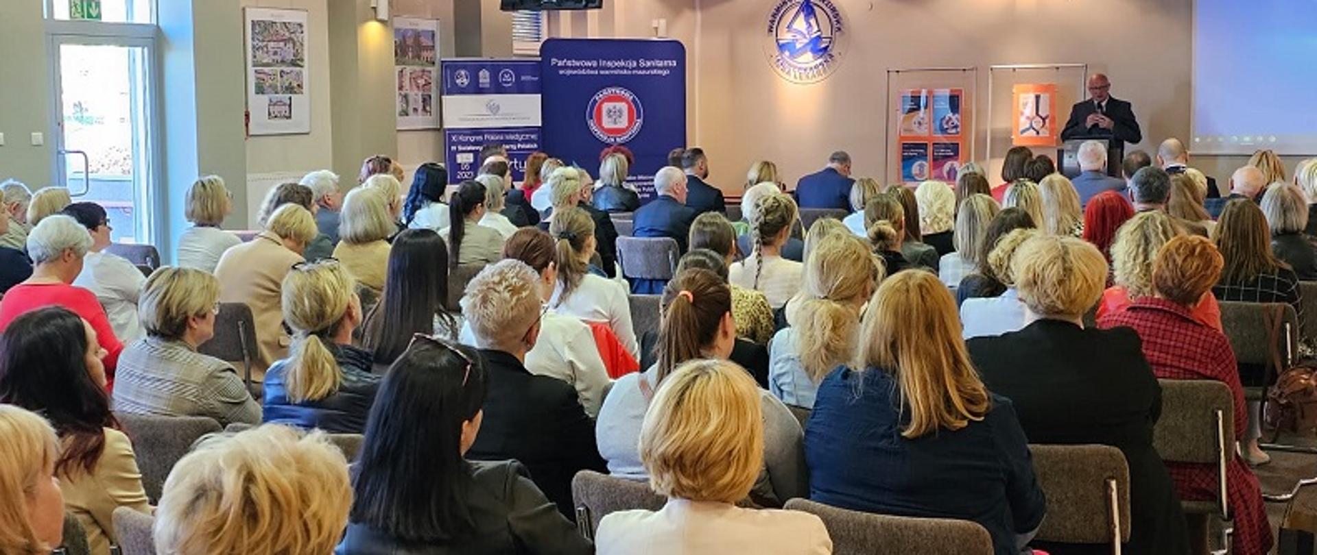 Przemawiający wojewódzki inspektor sanitarny oraz duża liczna uczestników siedzących na konferencji 