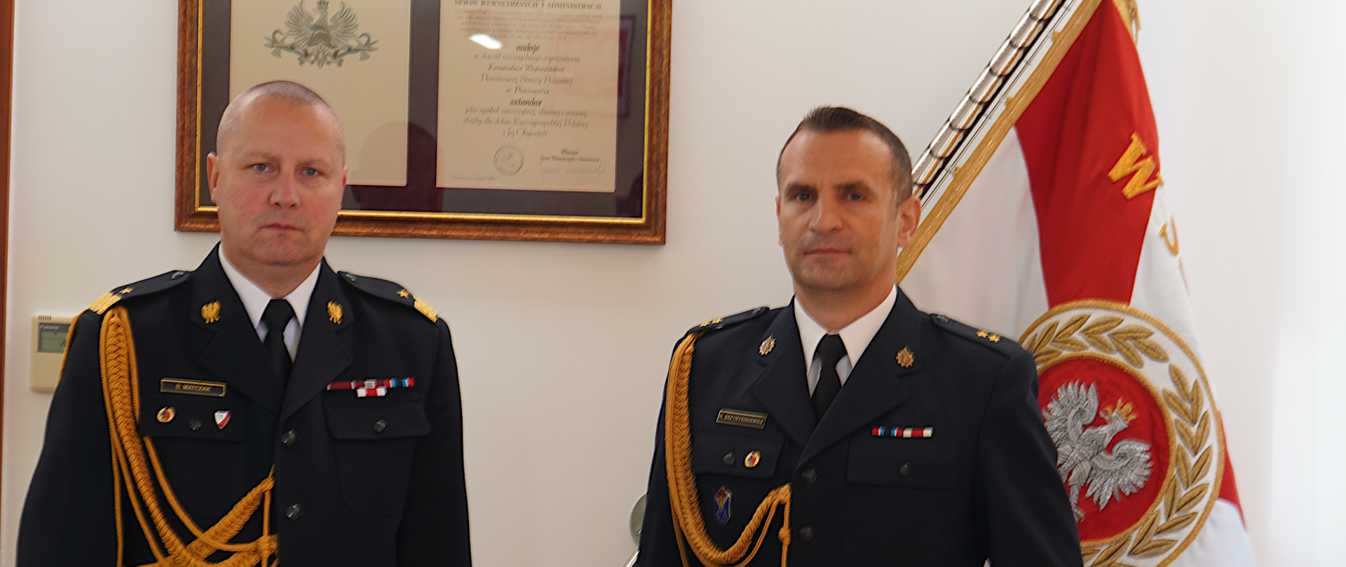 dwóch mężczyzn stoi obok siebie w ciemnych mundurach ze złotym sznurem i białych koszulach z czarnym krawatem. W tle sztandar