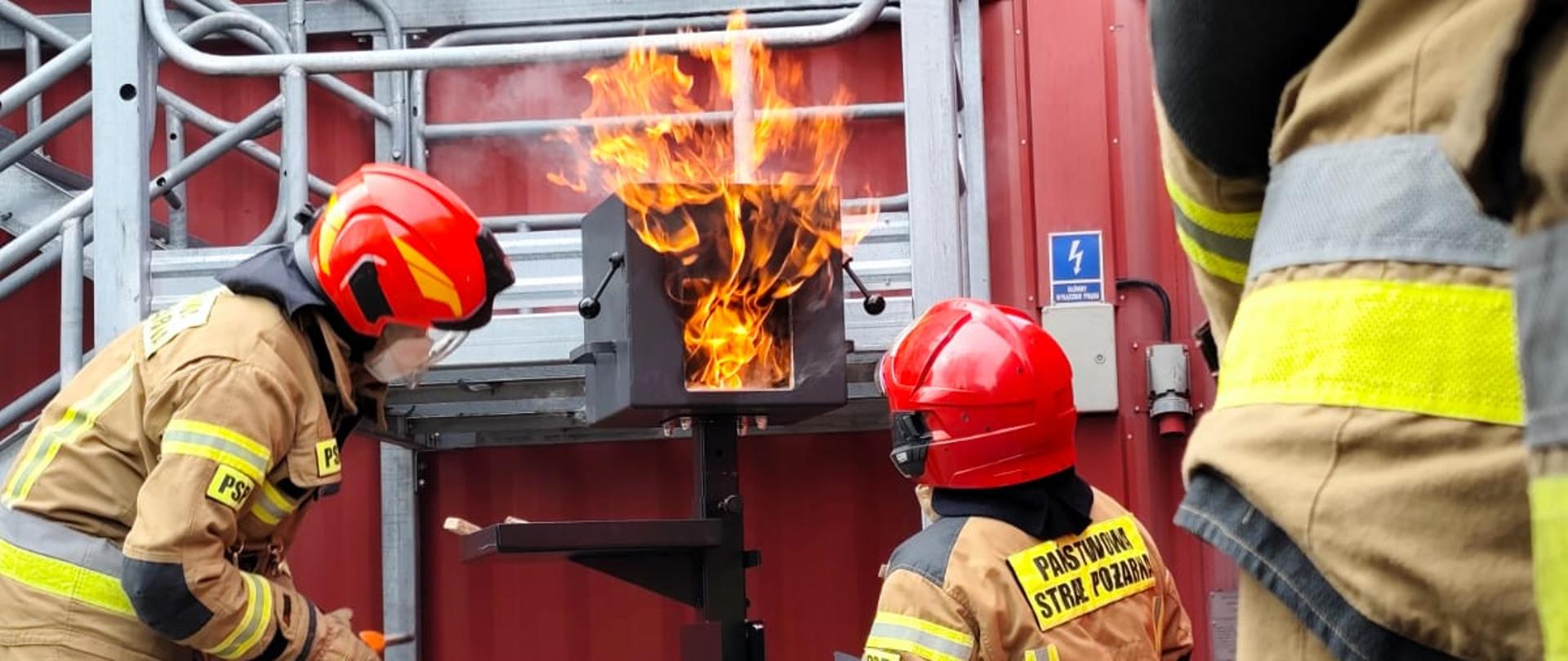 Bliski plan na dwóch strażaków w piaskowych mundurach bojowych i czerwonych hełmach PSP. Jeden strażak w kucki obsługuje mały, metalowy trenażer pożarowy, z którego bucha jasny ogień. Drugi strażak stoi obok. W tle widać metalowe konstrukcje schodów i trenażerów, w tym element czerwonego kontenera z tabliczką ostrzegawczą.