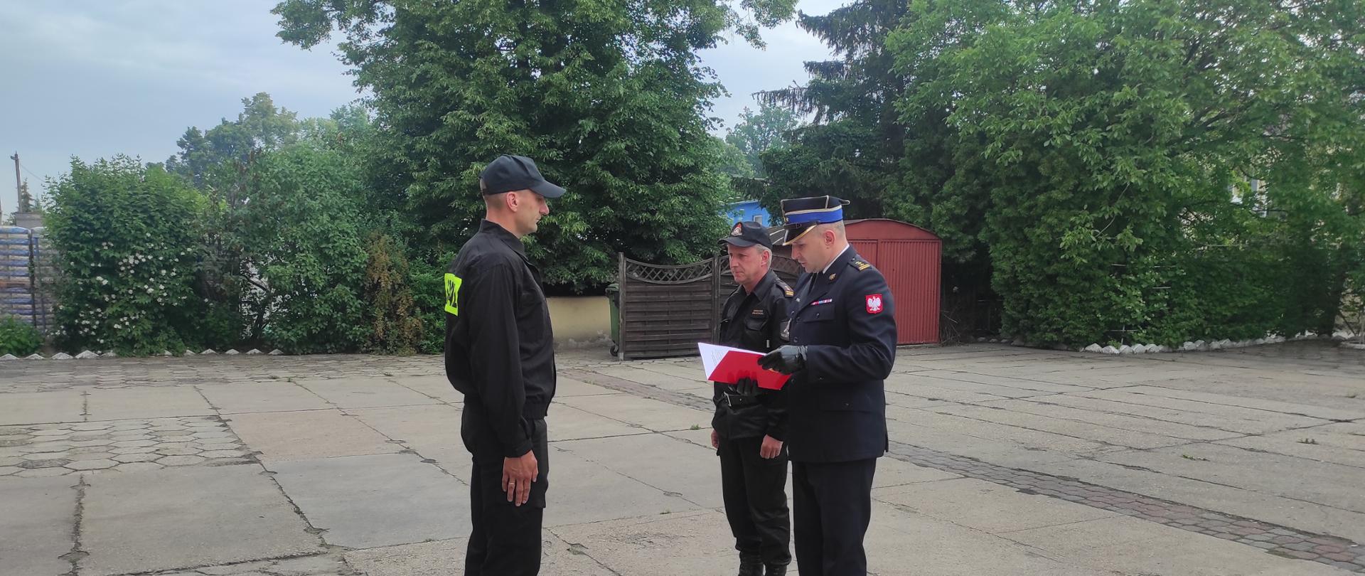 Na zdjęciu funkcjonariusze PSP przed budynkiem JRG 2 w Kaliszu. Strażacy umundurowani są w mundur koszarowy, KDS oraz wyjściowy. Zdjęcie w ciągu dnia. Na drugim planie zieleń. Strażacy stoją na szarym betonie.