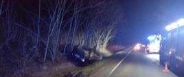 Zdjęcie wykonane w porze nocnej. Widać uszkodzony samochód w rowie. Na pierwszym planie płyta podłogowa. Światło sztuczne. W tle drzewa. W dolnej części zdjęcia droga publiczna.