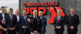 Strażak w mundurze wyjściowym, przed nim kilka mikrofonów, za nim kilka osób, stojących w grupie. W tle fragment budynku z czerwonym napisem.