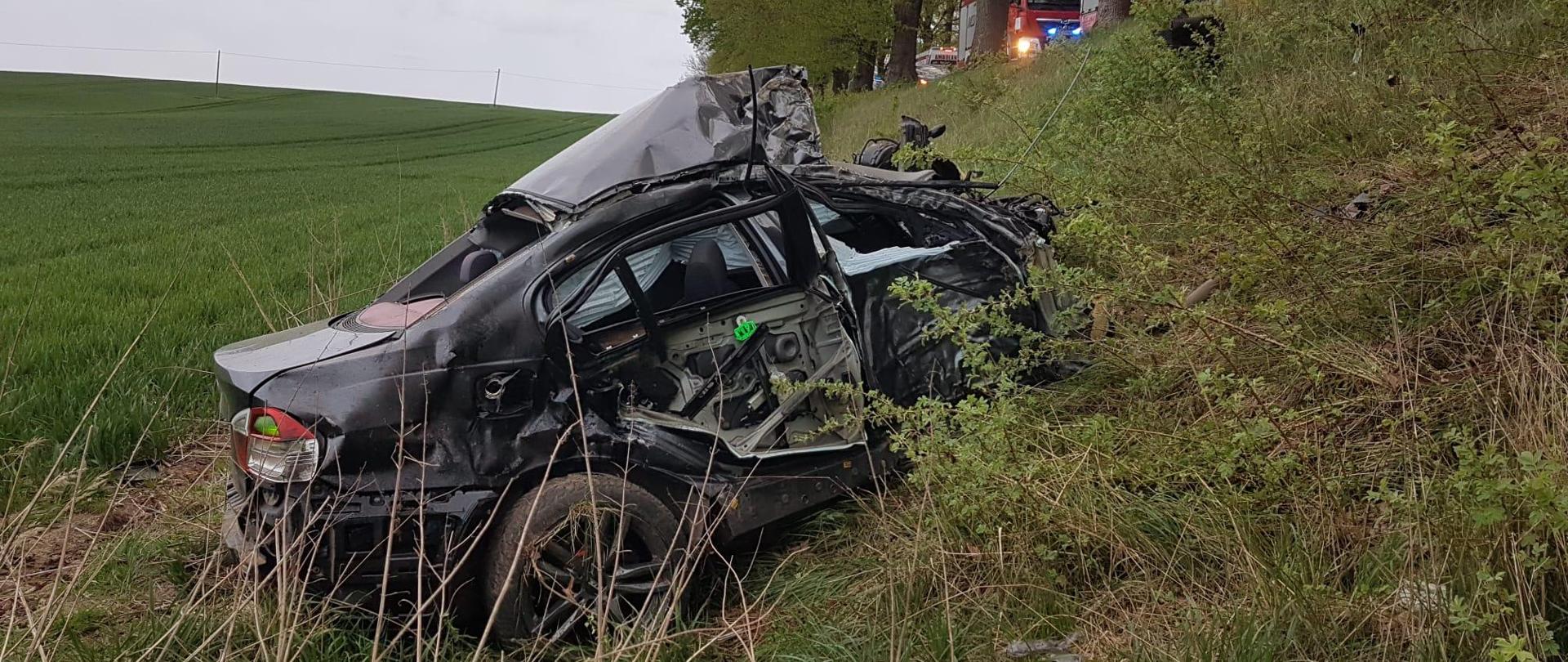 Na zdjęciu widoczny jest uszkodzony pojazd marki BMW. Auto ze zgniecionym przodem znajduje się w połowie przydrożnej skarpy. W tle, powyżej na jezdni ustawione są pojazdy ratownicze.