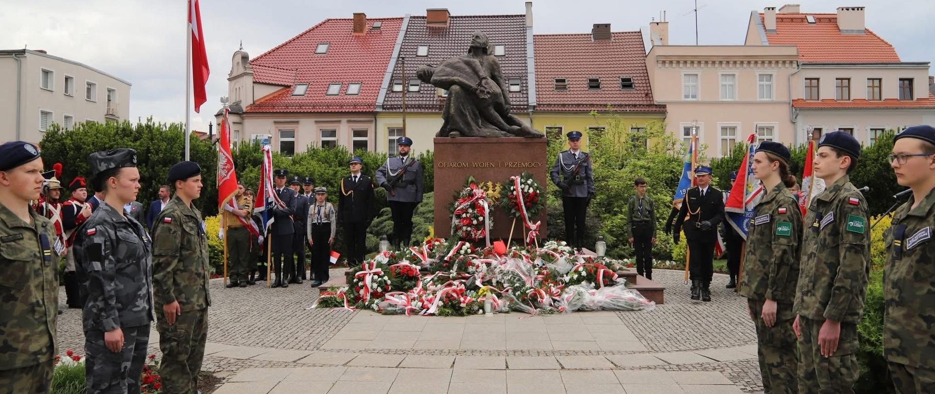 Na zdjęciu znajdują się służby mundurowe oraz uczniowie klas mundurowych w mundurach, pod pomnikiem złożone są wiązanki kwiatów. W tle widać budynki