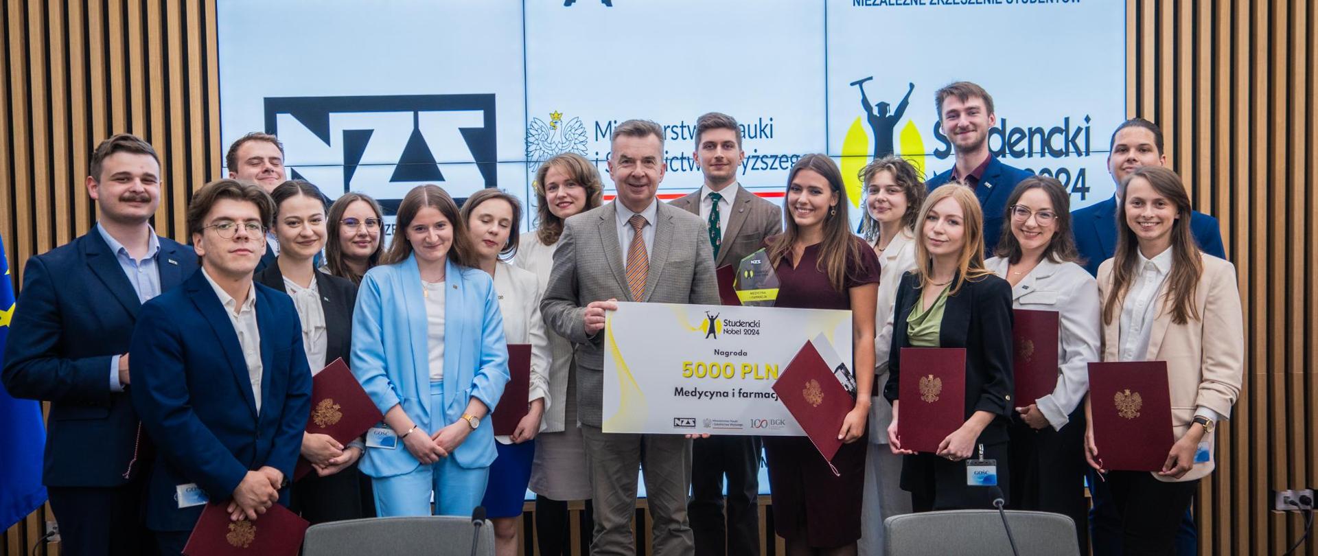 Zdjęcie zbiorowe, pod wbudowanym w ścianę wielkim ekranem stoi grupa młodych osób trzymających czerwone teczki, pośrodku grupy minister Wieczorek trzyma wielki symboliczny czek z napisem 5000 zł - Studencki nobel - medycyna i farmacja.