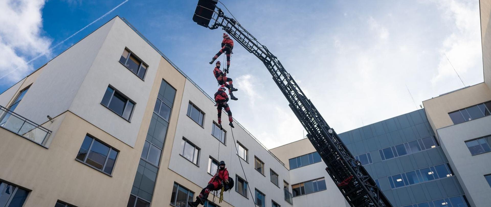 Na zdjęciu 4 ratownicy wysokościowi w czerwonych mundurach zjeżdżający na linkach alpinistycznych z kosza drabiny mechanicznej. W tle budynek szpitala.