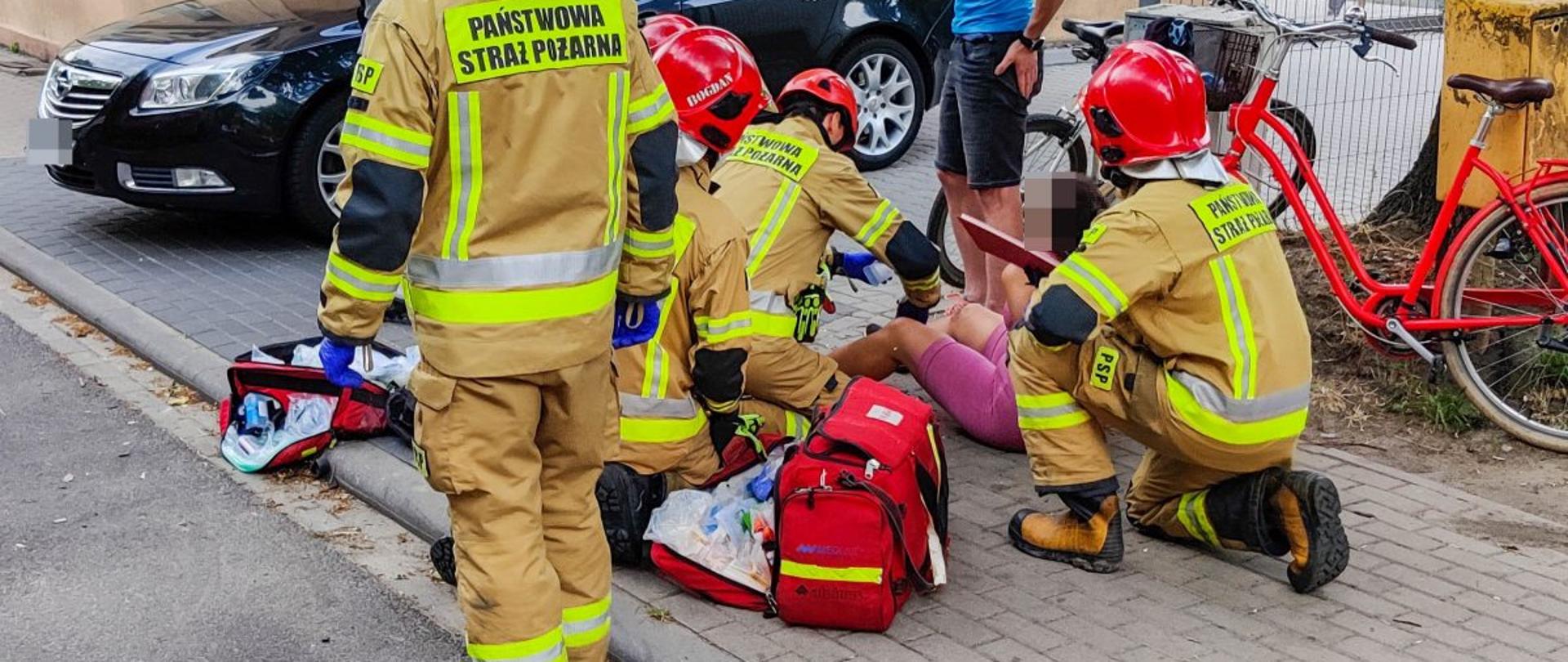 Na zdjęciu ratownicy PSP podczas udzielania osobie poszkodowanej - rowerzystce kwalifikowanej pierwszej pomocy, na chodniku pojazd osobowy oraz dwie osoby postronne, o drzewo oparty rower