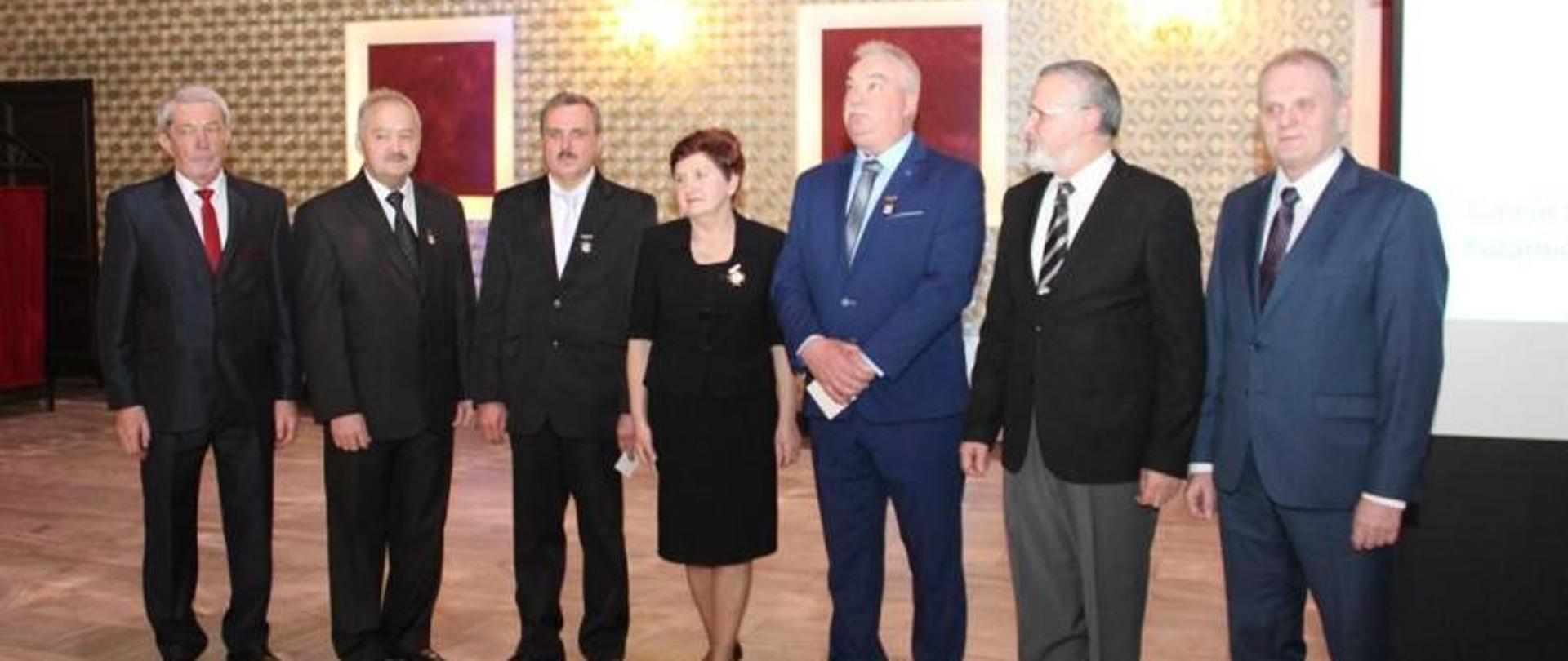 Na zdjęciu widzimy sześciu mężczyzn oraz kobietę stojących w szeregu i pozujących do zdjęcia. Kobieta znajduje się po środku. Mężczyźni ubrani wyjściowo w ciemne garnitury, jasne koszule oraz krawaty. Kobieta ubrana w czarną sukienkę do kolan. Za pozującymi widzimy wystrój pomieszczenia, zdobione ściany w kolorze złotym, kinkiety świecące. 