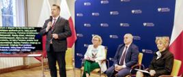 Minister Czarnek stoi i mówi do mikrofonu, za nim siedzą na krzesłach dwie kobiety i mężczyzna trzymający smycz do której przypięty jest leżący na podłodze czarny pies, za nimi granatowa ścianka.