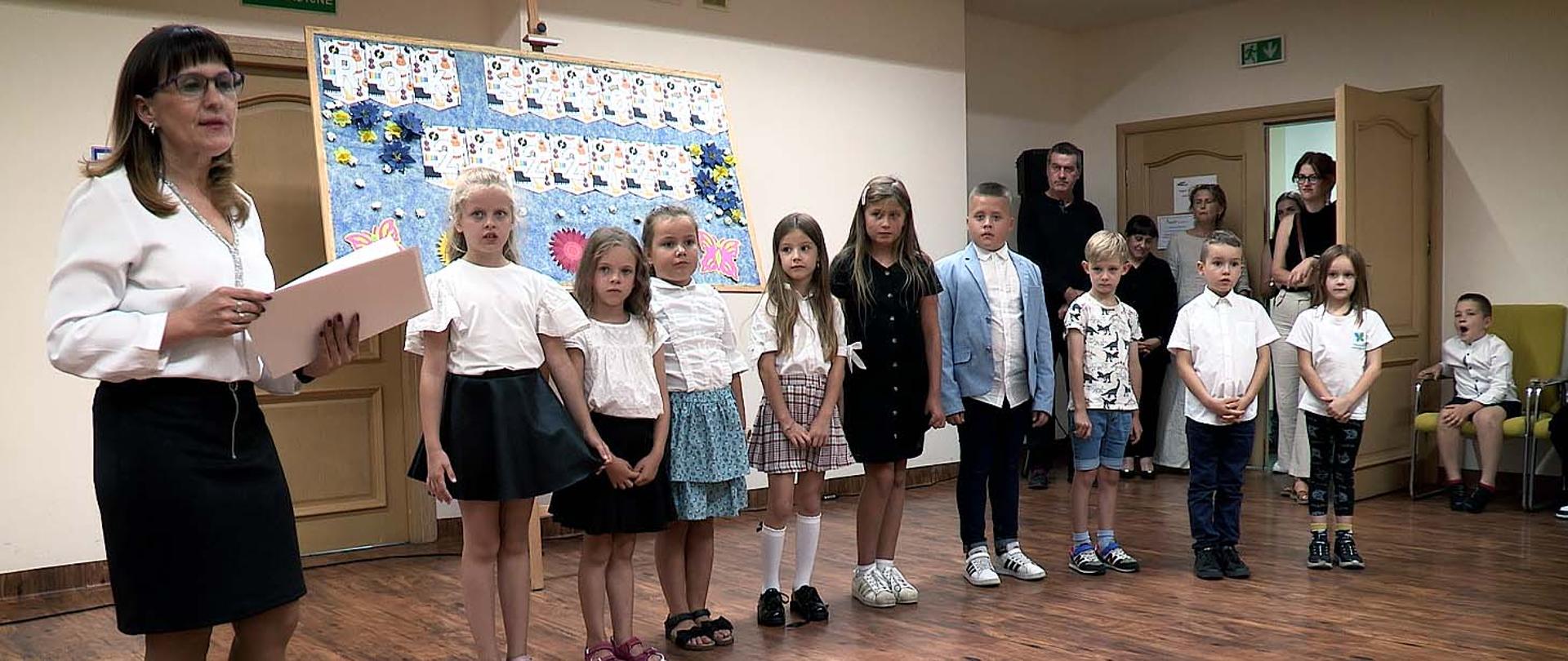 Na zdjęciu grupa osób stojąca w auli szkolnej. Z lewej strony nauczycielka, trzyma w ręku dokumenty, patrzy do przodu. Po jej lewej stronie stoi w rzędzie 9 uczniów skierowanych w przód, patrzących z boku na nauczycielkę. Z prawej strony zdjęcia, w drzwiach do sali stoją dorosłe osoby.