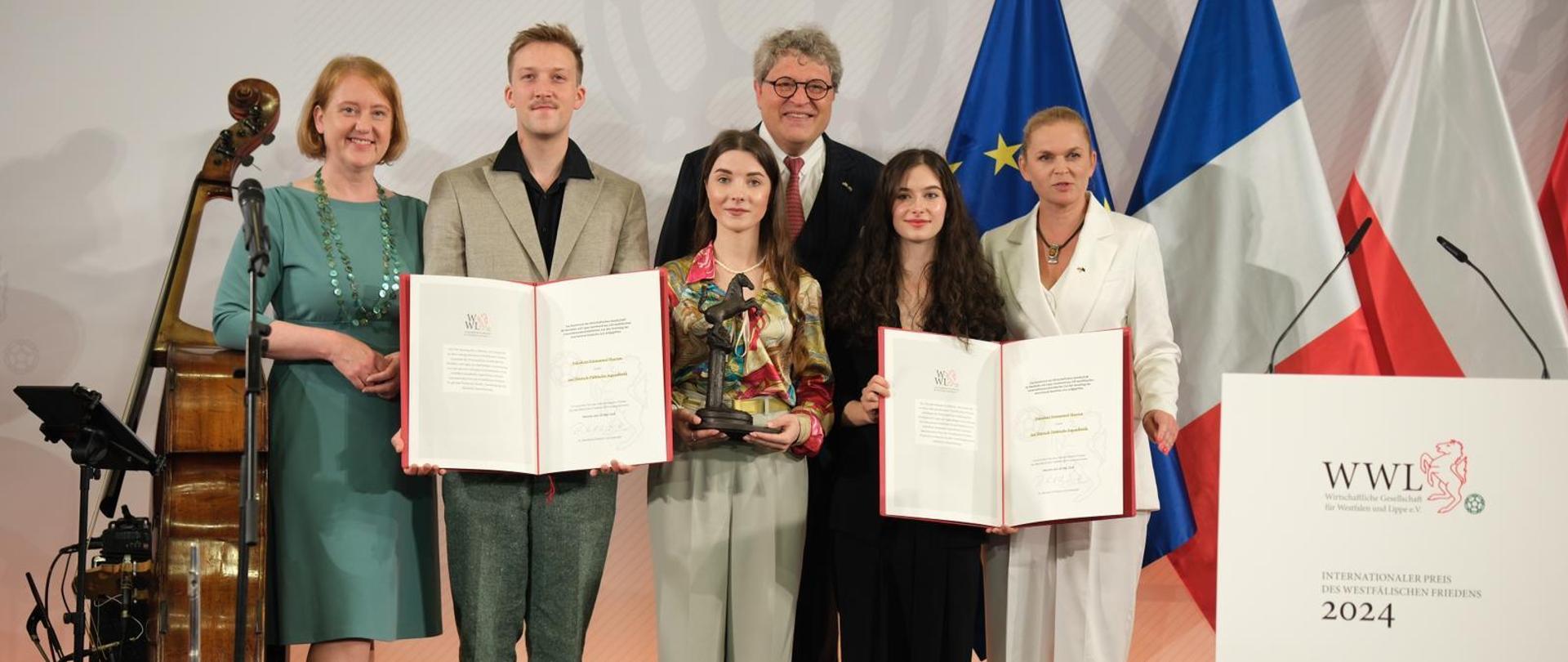 Minister Barbara Nowacka pozuje do zdjęcia z laureatami młodzieżowej Międzynarodowej Nagrody Pokoju Westfalskiego. Dwie osoby trzymają w ręku otwartą teczkę z dokumentem przyznającym nagrodę. Jedna osoba trzyma w ręku statuetkę przyznaną zwycięzcom. Za ich plecami stoją flagi Unii Europejskiej, Francji i Polski. 