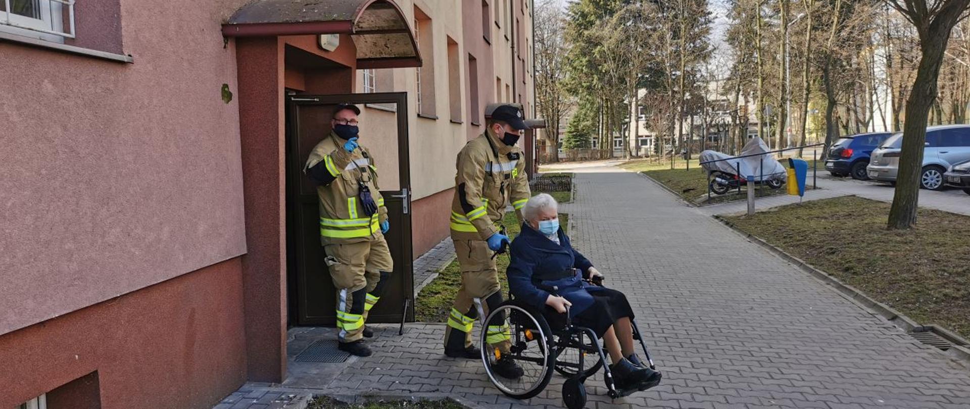 Zdjęcie przedstawia strażaków z OSP Osiedle Stałe wyprowadzających z bloku kobietę poruszającą się na wózku inwalidzkim w ramach akcji #SzczepimySię.