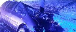 Na zdjęciu samochód osobowy po wypadku, znajdujący się w rowie obok przydrożnych drzew. Widoczny jest uszkodzony przód pojazdu. 