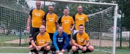 na zdjęciu zawodnicy na tle bramki 7 funkcjonariuszy reprezentujących KP PSP Śrem w koszulkach pomarańczowo - czarnych bramkarz w koszulce niebieskiej trzyma piłkę kuca 3 zawodników za nimi 4 stoi