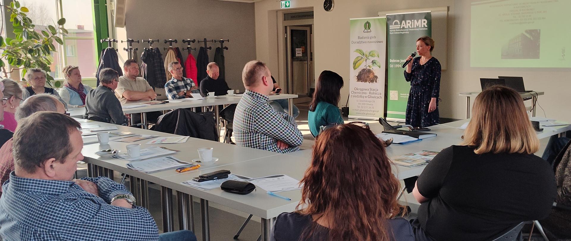 Na zdjęciu widać salę szkoleniową, w której odbywa się spotkanie lub prezentacja. Uczestnicy – około kilkanaście osób, głównie dorośli – siedzą przy ustawionych w prostokąt stołach. Na stołach leżą dokumenty, długopisy, telefony oraz filiżanki z kawą lub herbatą. Większość osób jest zwrócona w stronę prowadzącej i słucha uważnie, niektórzy mają skrzyżowane ręce lub notują.
Po prawej stronie stoi kobieta prowadząca spotkanie. Trzyma mikrofon i przemawia do grupy. Za nią znajduje się ekran z wyświetloną prezentacją oraz dwa stojące banery informacyjne. Obok stoi stolik z laptopem podłączonym do projektora.
Pomieszczenie jest jasne, oświetlone światłem dziennym wpadającym przez duże okno po lewej stronie. Przy oknie stoi duża roślina doniczkowa, a pod ścianą znajdują się wieszaki z powieszonymi kurtkami uczestników.
Atmosfera wygląda na spokojną i skupioną – to formalne szkolenie lub spotkanie informacyjne.