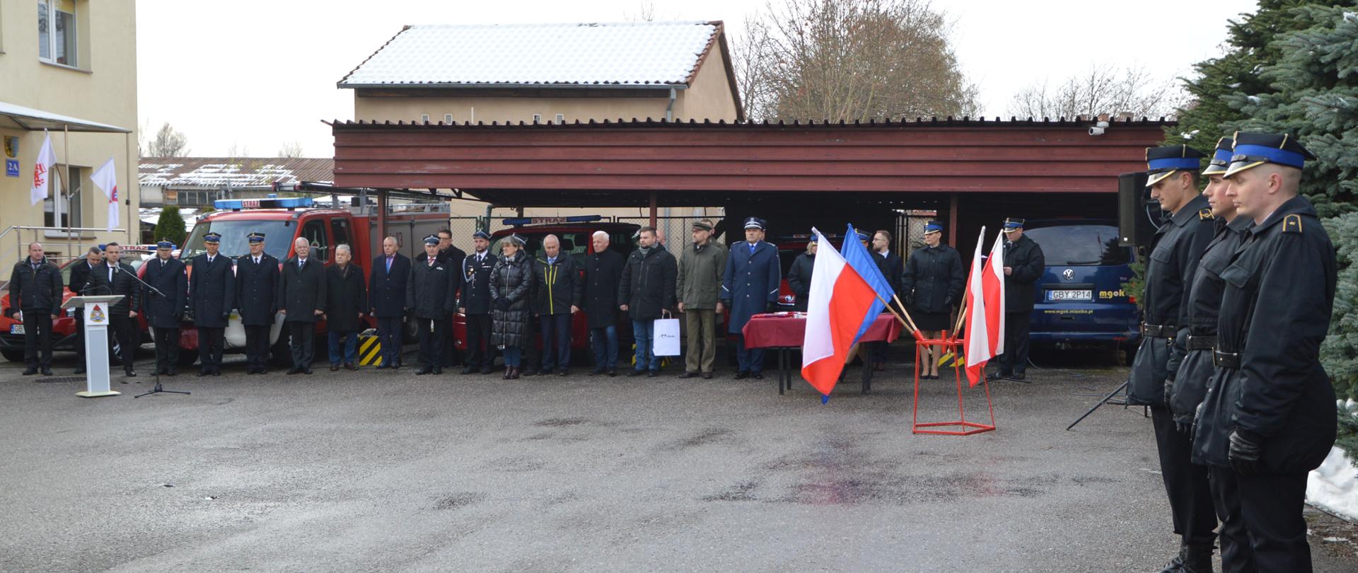 Po prawej stronie widać trzech strażaków ubranych w mundury wyjściowe koloru czarnego. W oddali na tle samochodów widać grupę zaproszonych gości.