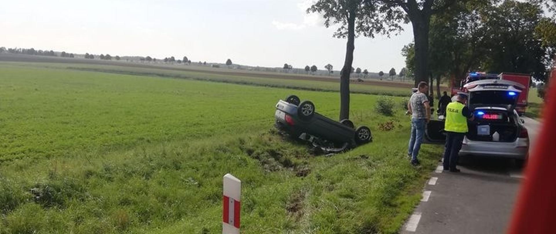 Samochód osobowy leżący na dachu w rowie. Policjant spisuje dane.