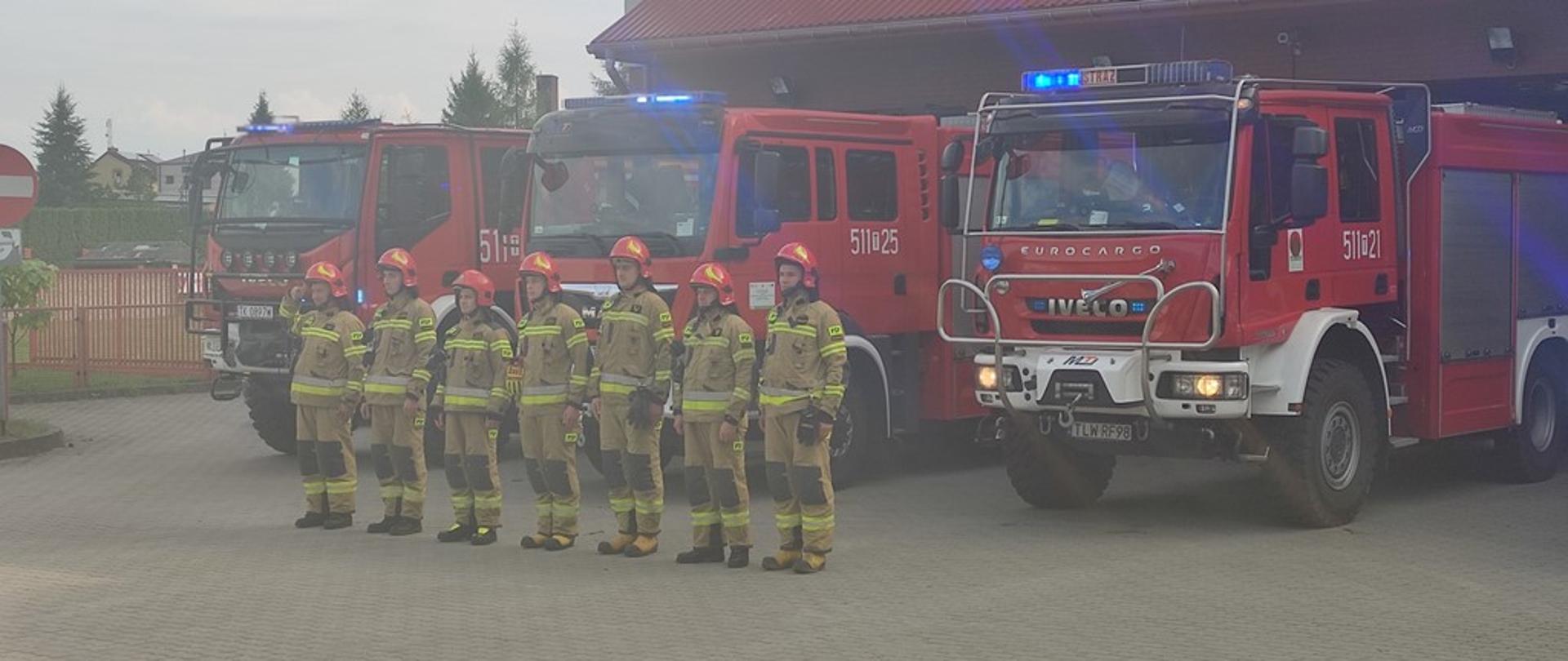 Przed budynkiem KP PSP we Włoszczowie, strażacy podziału bojowego w ubraniach specjalnych, ustawieni w szeregu, pojazdy pożarniczej mają włączone sygnały dźwiękowe i świetlne
