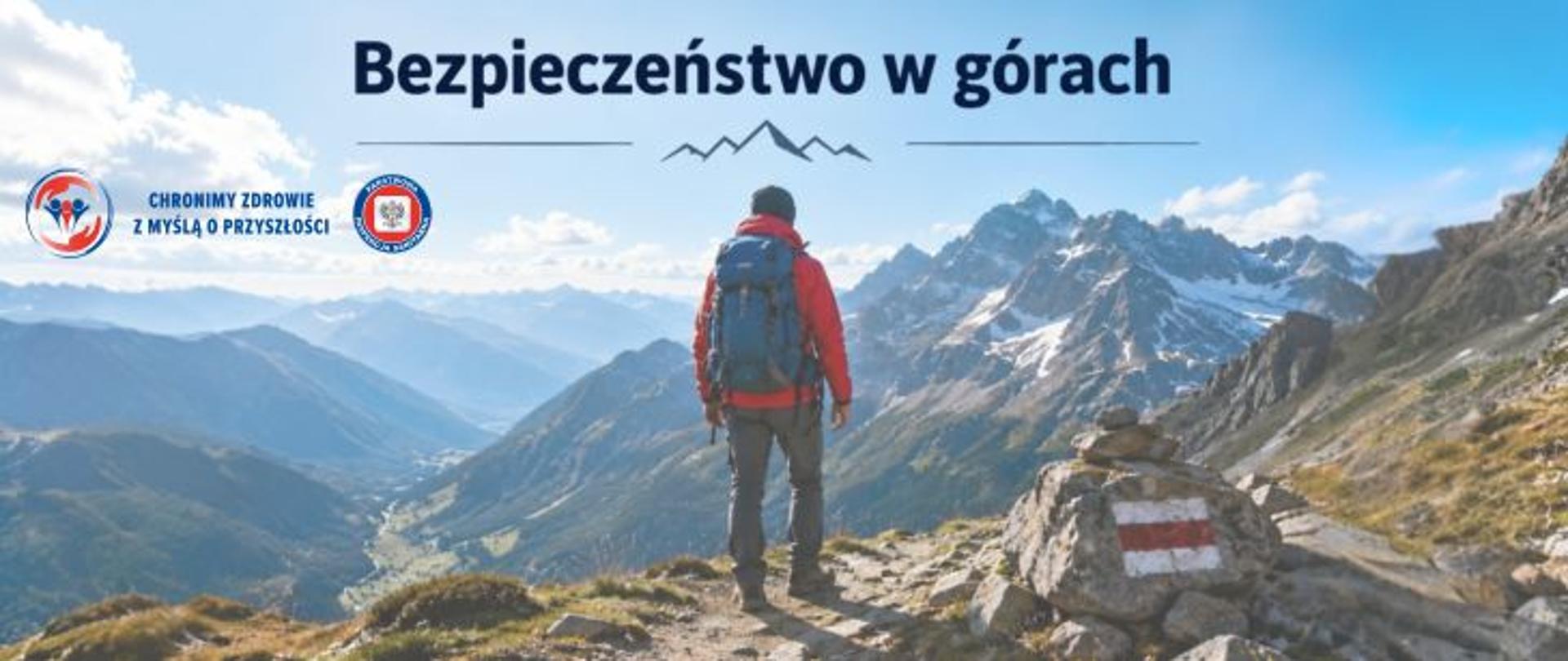 Baner przedstawiający turystę w czerwonej kurtce z niebieskim plecakiem stojącego na górskim szlaku i podziwiającego panoramę ośnieżonych szczytów. U góry znajduje się tytuł „Bezpieczeństwo w górach”. Na dole po prawej stronie widoczne oznaczenie szlaku w kolorach białym i czerwonym. Po lewej stronie logo Państwowej Inspekcji Sanitarnej wraz z hasłem: Chronimy zdrowie z myślą o przyszłości. 
