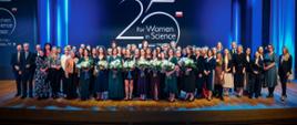 Na tle granatowej ściany z napisem 25 for Women in Science stoi kilkadziesiąt osób, głównie elegancko ubranych kobiet, kobiety z przodu trzymają bukiety białych kwiatów.