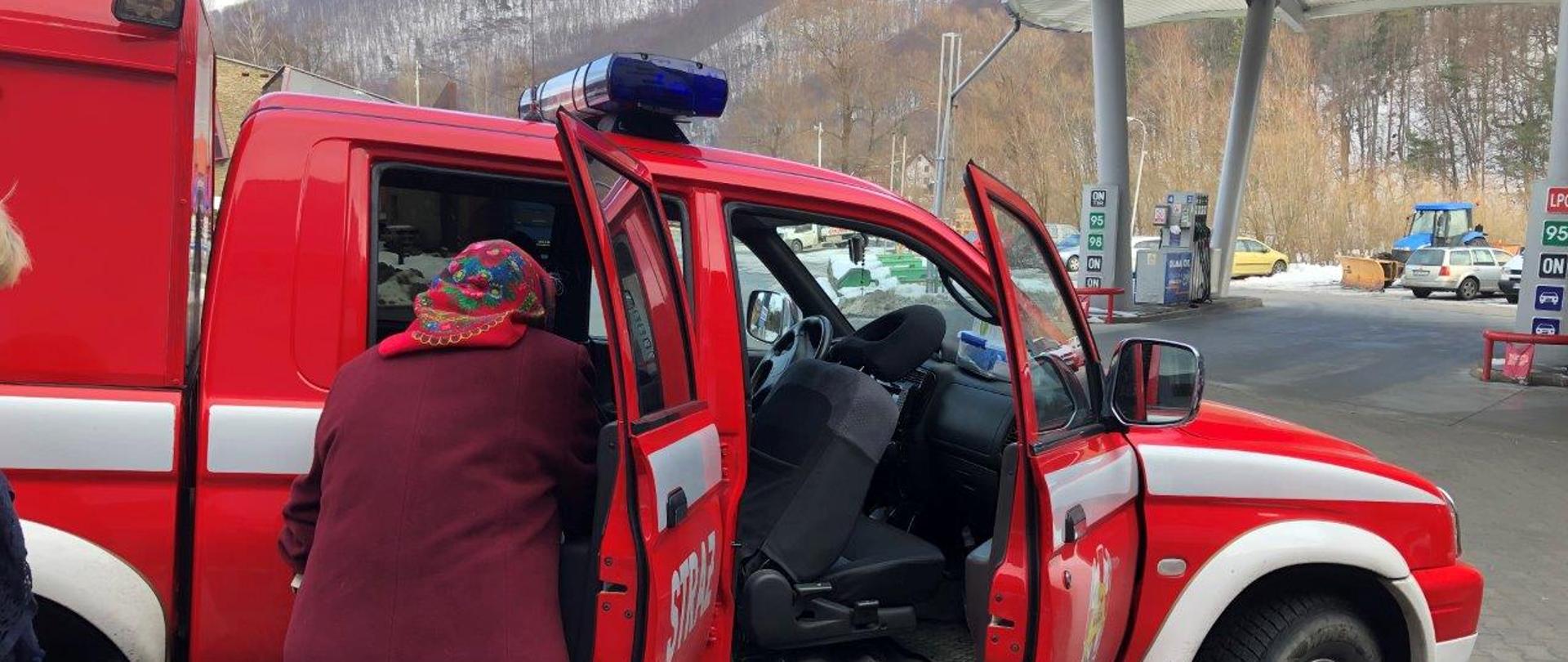 Starsza kobieta w bordowym płaszczu i chustce na głowie wsiada do samochodu PSP