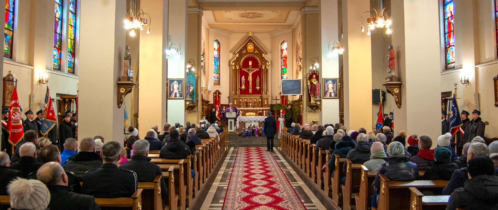 Uczestniczy mszy świętej siedzą w ławkach i słuchają kazania głoszonego przez księdza stojącego przy ołtarzu. W nawie centralnej widoczny dowódca uroczystości stojący na baczność, a po nawach zewnętrznych kościoła poczty sztandarowe 