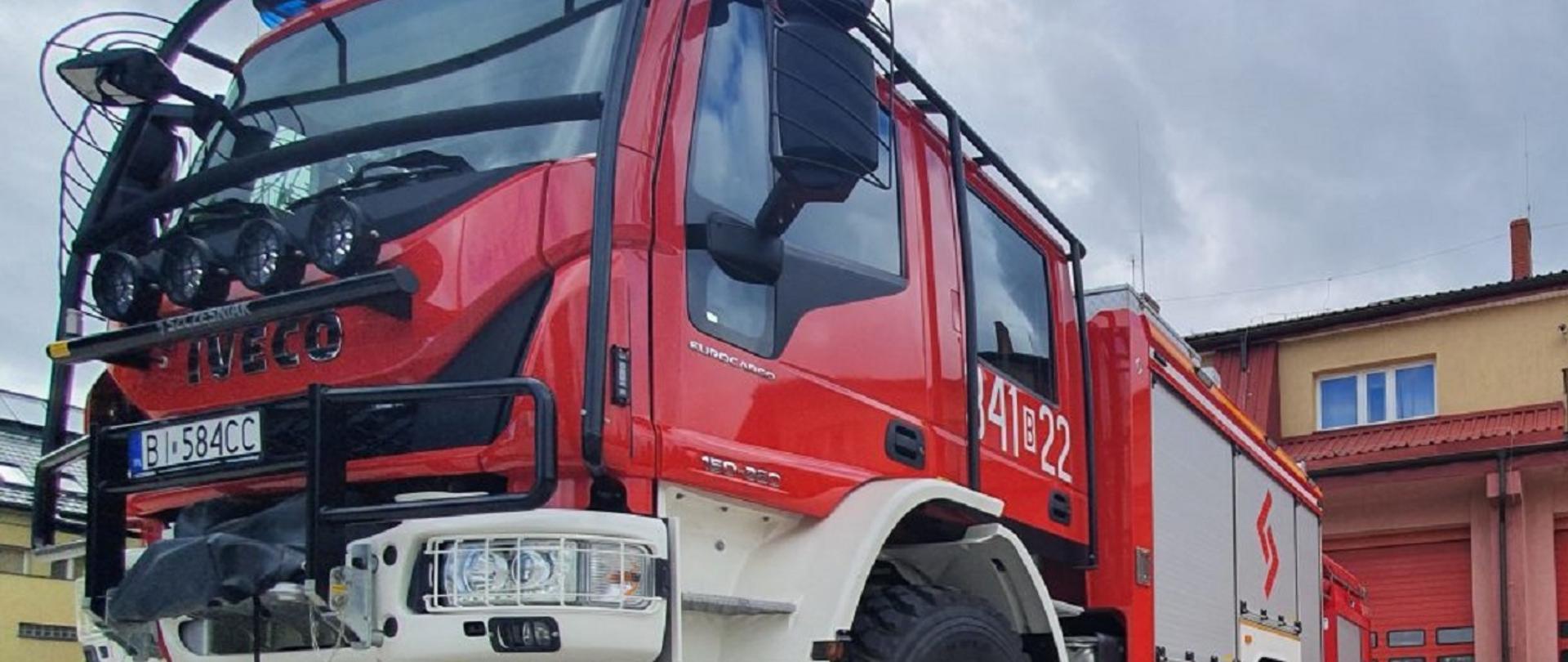Samochód średni gaśniczy w kolorze czerwony z białymi zderzakami. W drugim planie zdjęcia dwa budynki. Pierwszy budynek Jednostki Ratowniczo Gaśniczej numer 1 z bramami garażowymi koloru czerwonego. Drugi budynek w kolorze żółtym Stacji Ratownictwa Medycznego.