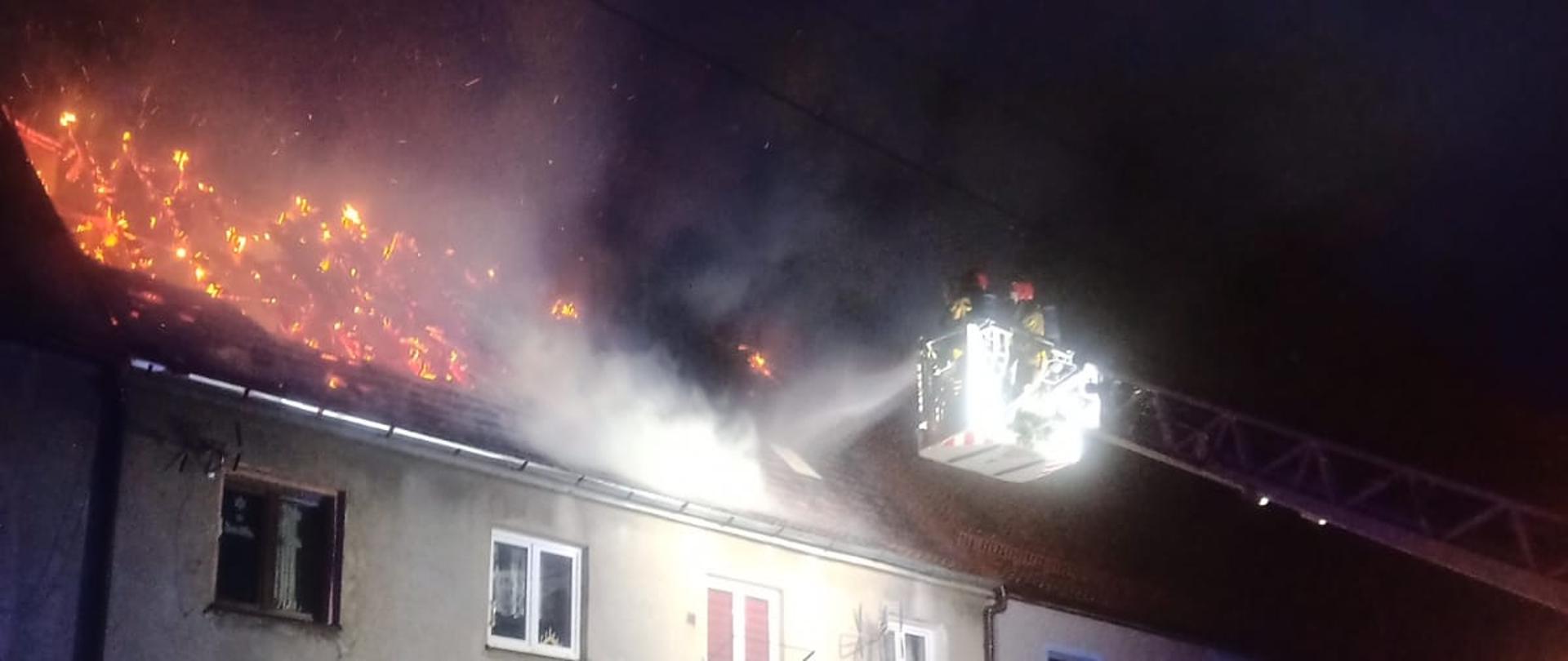 Strażacy gaszą wodą z działka drabiny mechanicznej pożar dachu w budynku mieszkalnym wielorodzinnym