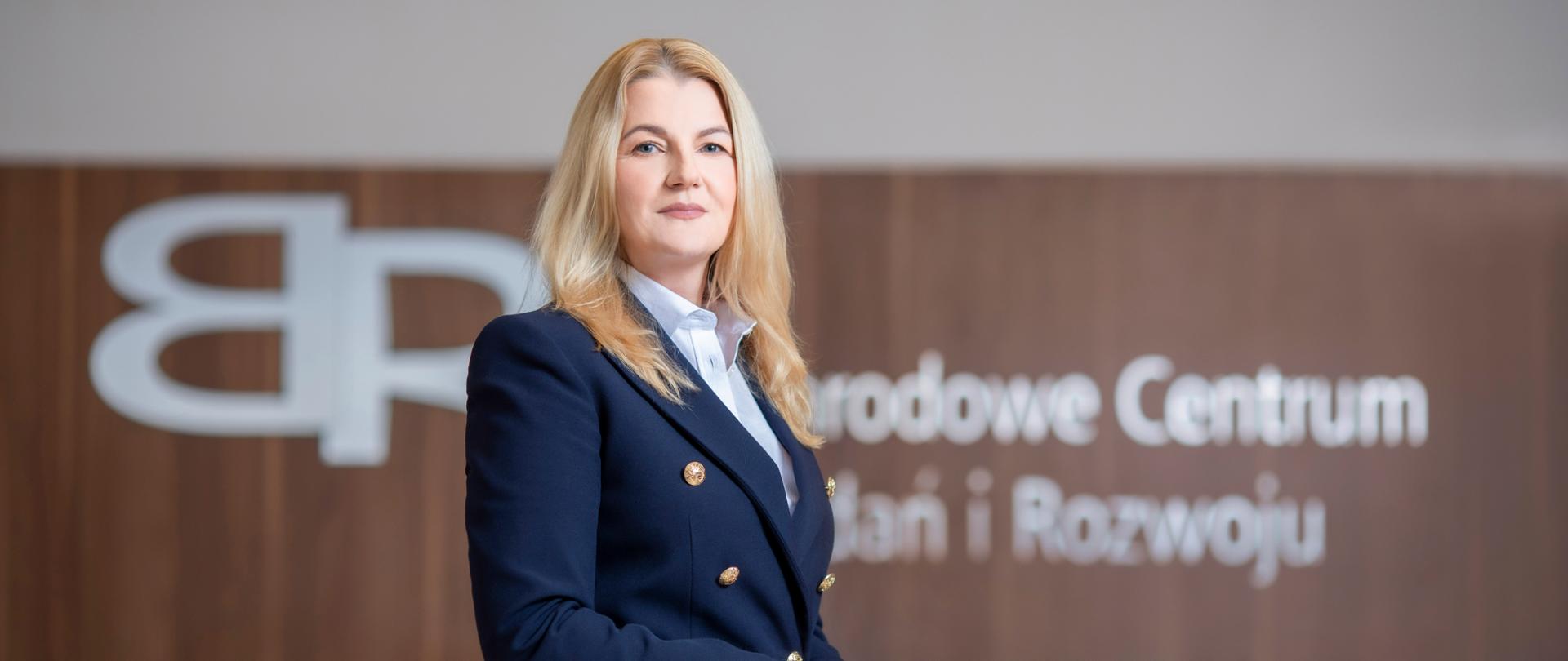 Against the background of the inscription National Center for Research and Development, a woman standing sideways in a navy blue jacket