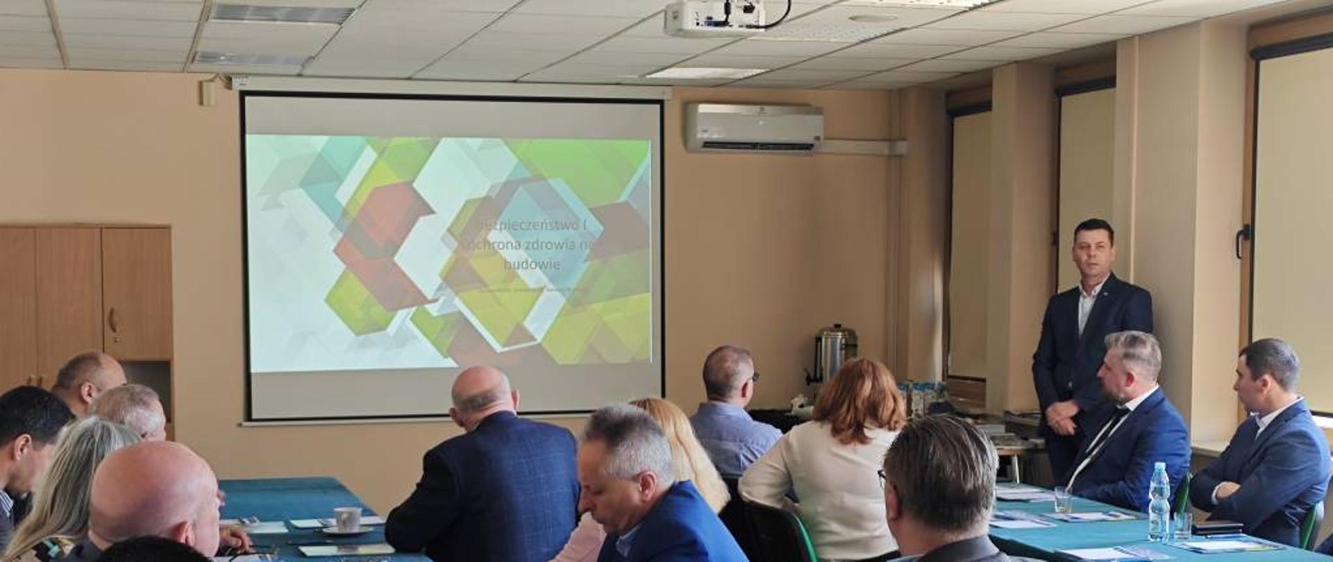 W sali znajduje się grupa osób siedzących przy stołach, zaangażowanych w rozmowy i interakcje. 