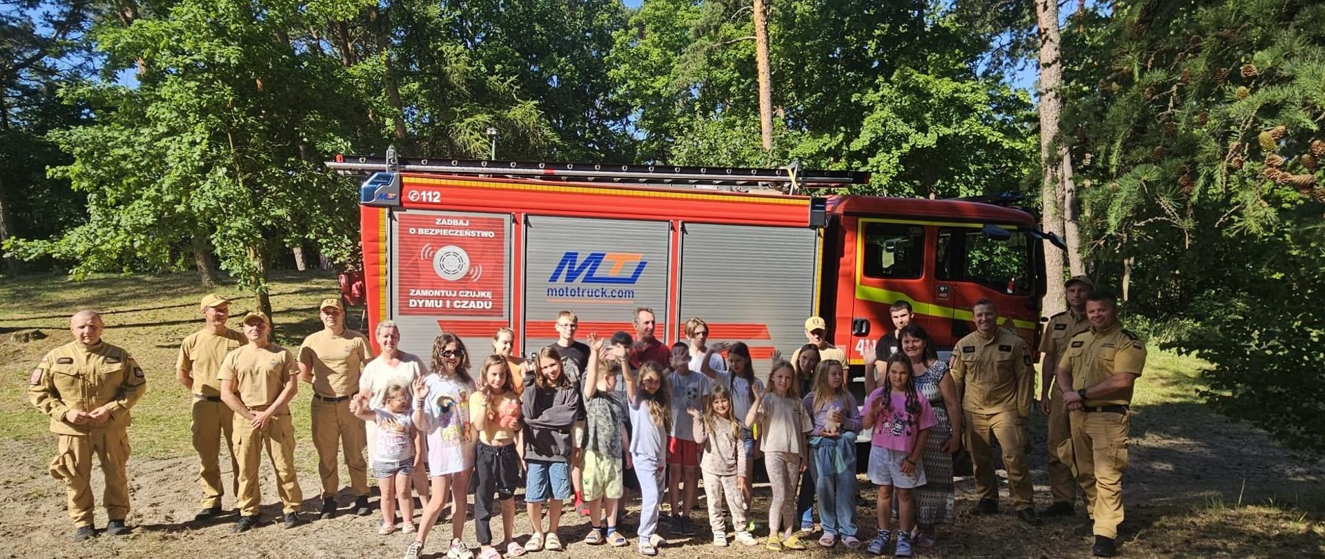Strażacy z harcerzami na tle samochodu strażackiego na leśnej polanie. Słonecznie.