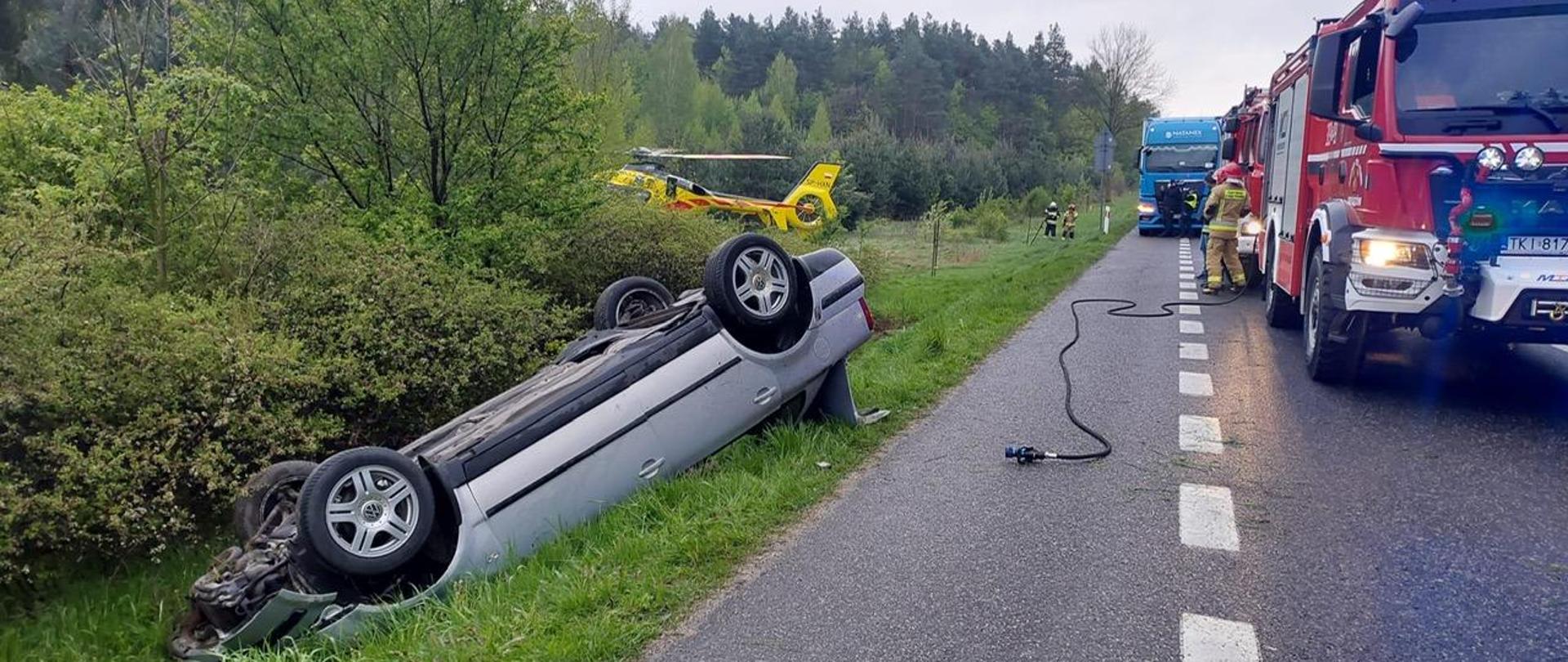 Zdjęcie przedstawia drogę krajową. Po lewej stronie, na poboczu, leży na dachu samochód osobowy. Po prawej stronie, na jezdni stoją dwa auta strażackie, a w głębi widać fragment ratowniczego śmigłowca.