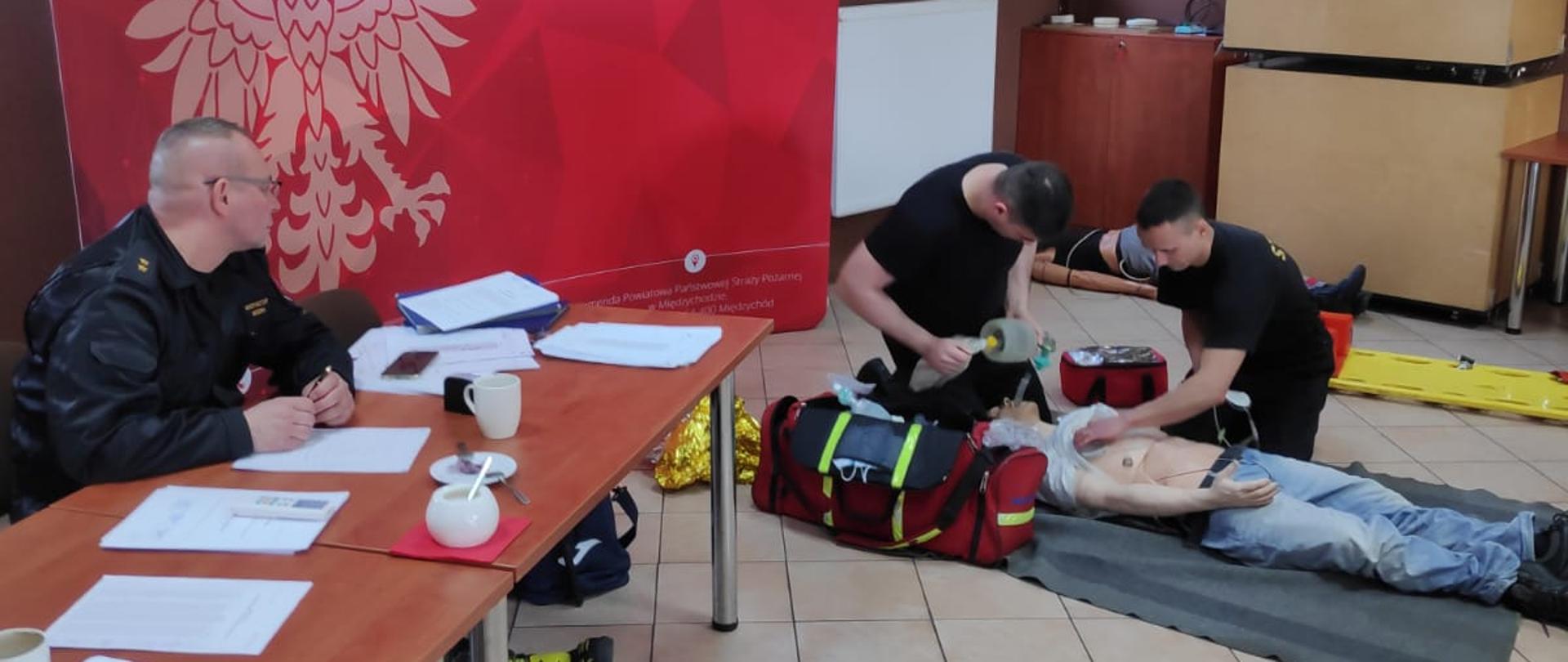 Na zdjęciu widać egzaminatora oraz dwóch funkcjonariuszy udzielających pierwszej pomocy na fantomie ćwiczebnym podczas egzaminu