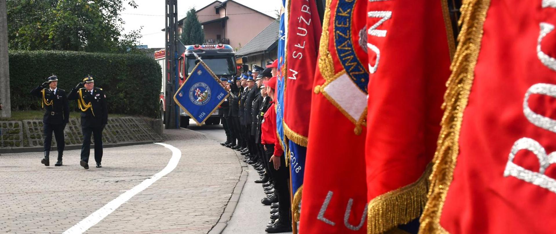 Zdjęcie przedstawia uroczystą paradę lub ceremonię straży pożarnej w miejscowości Przysucha. Na pierwszym planie po prawej stronie widać szereg sztandarów z frędzlami, w tym co najmniej trzy czerwone i jeden niebieski, z widocznymi napisami, takimi jak "W SŁUŻB..." i "ZWIĄZEK". Po lewej stronie zdjęcia, na wybrukowanej ulicy, dwaj mężczyźni w galowych mundurach strażackich salutują. Dalej w tle, wzdłuż ulicy, stoi rząd strażaków w mundurach, a za nimi widoczny jest wóz strażacki z częściowo zasłoniętą rejestracją oraz budynki mieszkalne i zieleń. Całość sugeruje oficjalne wydarzenie o charakterze patriotycznym lub honorowym.