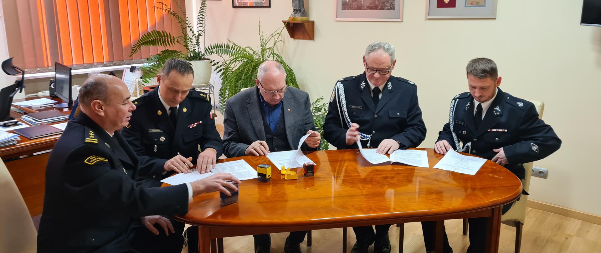 Czterech mężczyzn ubranych w czarne ubrania wyjściowe, sznury galowe, białe koszule, czarnym butami, siedzą przy stole. Jeden mężczyzna ubrany w ciemna marynarkę i ciemną koszulę siedzi przy stole. Mężczyźni siedzą i podpisują dokumenty. Na stole znajdują się dokumenty, woda mineralna . W tle na ścianie znajduje się obrazy oraz żaluzje koloru brązowego