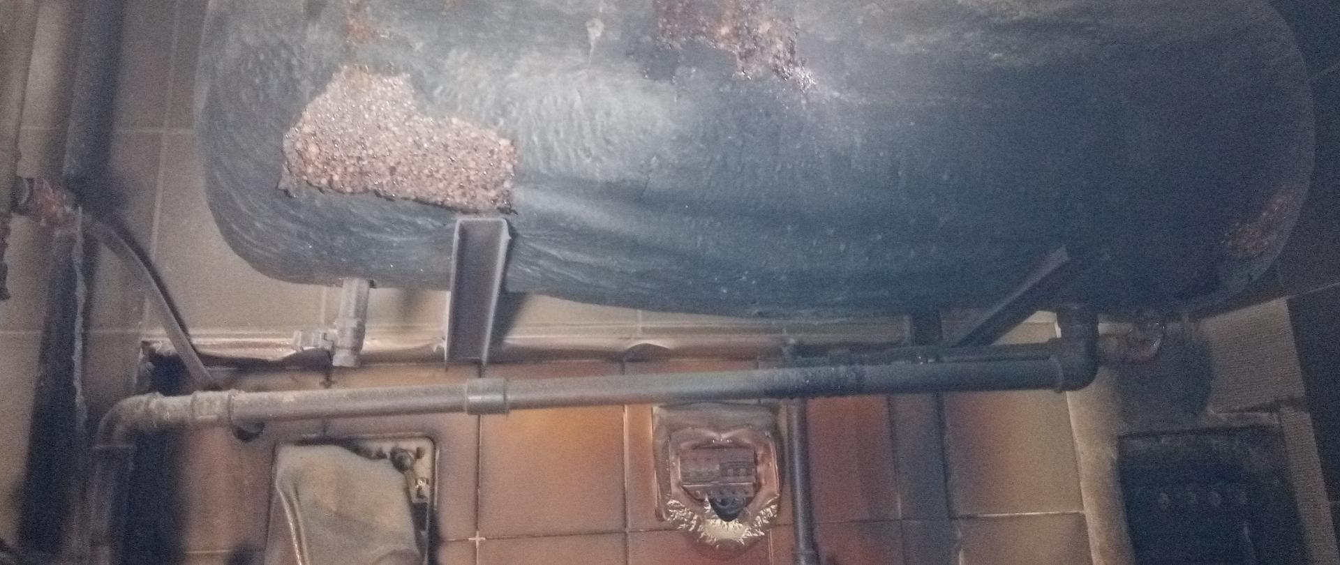 Pomieszczenie kotłowni. Zniszczony w wyniku pożaru bojer. Okopcona ściana, stopione elementy instalacji użytkowych na ścianie.