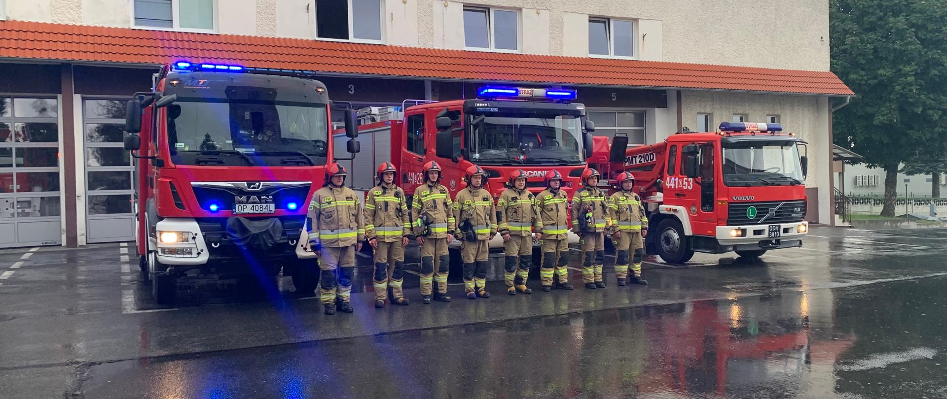 Na tle budynku JRG ustawiono trzy samochody pożarnicze: od lewej GBA, GCBA i podnośnik. Samochody małą włączone światła alarmowe. Przed samochodami w szeregu, w pozycji zasadniczej ustawieni są strażacy PSP (8) w piaskowych ubraniach specjalnych i czerwonych hełmach. 