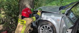 Samochód osobowy uderzył w drzewo, przy samochodzie jest strażak. 