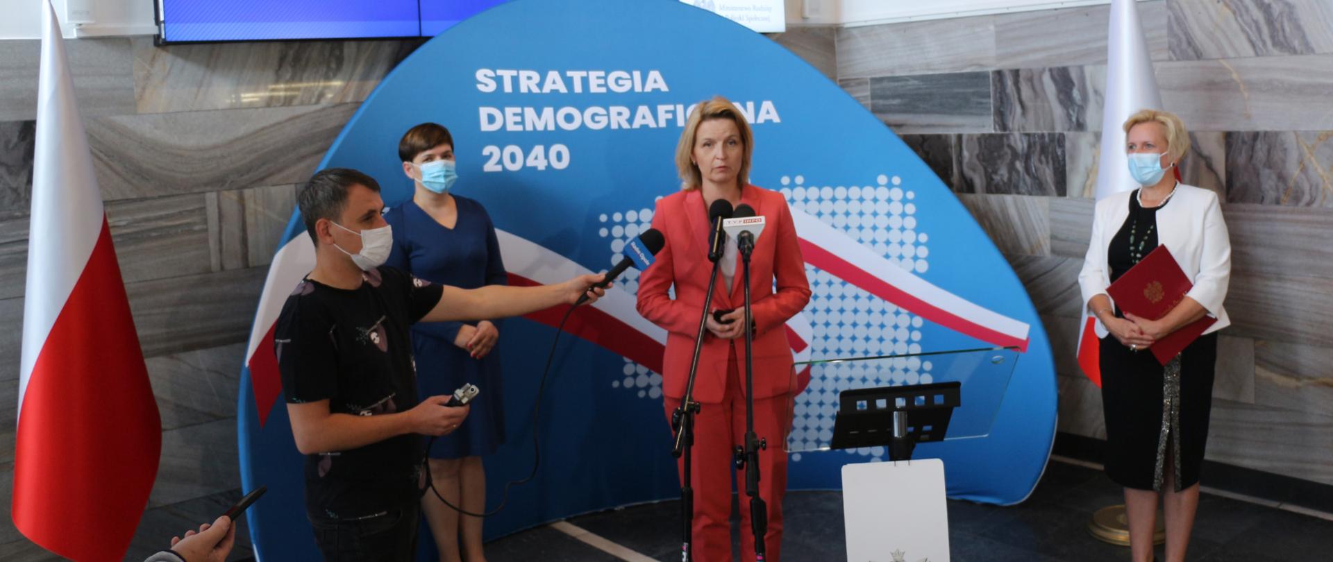 Zdjęcie z konferencji prasowej. W centralnej części fotografii widać ściankę z napisem Strategia Demograficzna 2040. To półokrągła ścianka w kolorze niebieskim z napisami w kolorze białym oraz biało-czerwoną wstęgą. Przed ścianką na środku stoi wiceminister rodziny i polityki społecznej. To kobieta ubrana w czerwony żakiet i beżową bluzkę. Po lewej stronie, za plecami minister, widać stojącą na tle ścianki kobietę, ubraną w niebieską sukienkę. Na przeciw kobiety widać dziennikarza, który stoi w bliskiej odległości od minister. W swoich dłoniach trzyma mikrofon. Po prawej stronie kadru widać wicewojewodę opolskiego, ubraną w czarną sukienkę i biały żakiet. Wicewojewoda spogląda na dziennikarza, który stoi obok minister. Po obu stronach kadru widać biało-czerwone flagi na stojakach. W tle szara, marmurowa ściana, jeden z elementów wnętrza holu urzędu. Na samej górze kadru widac monitor, na którym wyświetla siegranatowa planasza z napisem Strategia Demograficzna 2040.