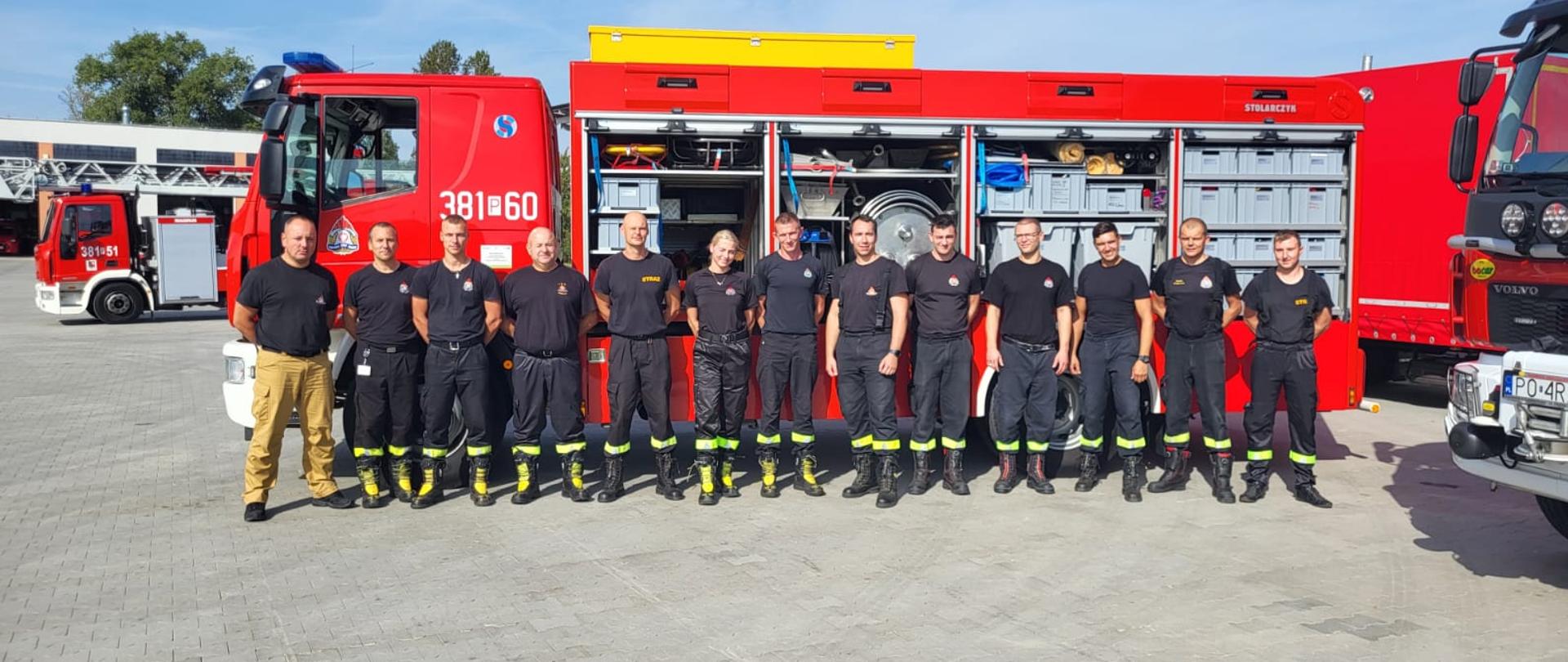 Strażacy stoją w szeregu na tle pojazdów strażackich.